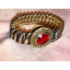 May include: A vintage gold-tone bracelet with a large, oval, red gemstone centerpiece. The bracelet features an ornate design with a stretchable, linked band. The gemstone is set in a decorative, filigree-style setting.