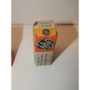 May include: A vintage General Electric vacuum tube box. The box is orange and grey, featuring the GE logo. Text on the box reads "2HG7/12GN7" and "Progress is Our Most Important Product."