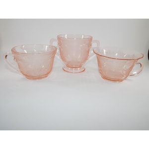 Puede incluir: Tres piezas de cristalería rosa, que incluyen dos tazas con asas y un azucarero. La cristalería presenta un estampado floral y está colocada sobre un fondo blanco. Las piezas están hechas de un material translúcido.