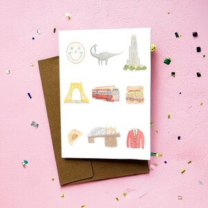 May include: A white greeting card with watercolor illustrations of a smiley face, dinosaur, building, bridge, sandwich, and red jacket. The card is on a brown envelope, with a pink background and confetti.