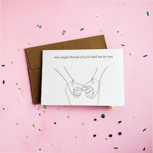 May include: A white greeting card with the text "one single thread of gold tied me to you" and an illustration of two hands holding pinky fingers. A brown envelope is behind the card, with confetti on a pink background.