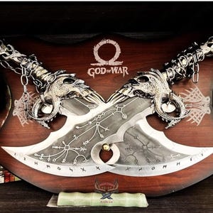 Könnte beinhalten: Zwei silberne, dekorative Schwerter mit aufwendigen Designs, montiert auf einer Holzplatte mit den Worten "God of War" und einem stilisierten Omega-Symbol.