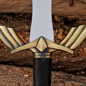 May include: Close-up of a sword with a bright white blade and a gold-colored hilt. The hilt features a wing-like design and a black, textured grip. The sword is set against a natural wood background.