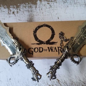 以下が含まれることがあります： 黒い柄のついた、銀色の装飾的な剣のペア。剣は、「God of War」と書かれた茶色の箱の上に置かれています。