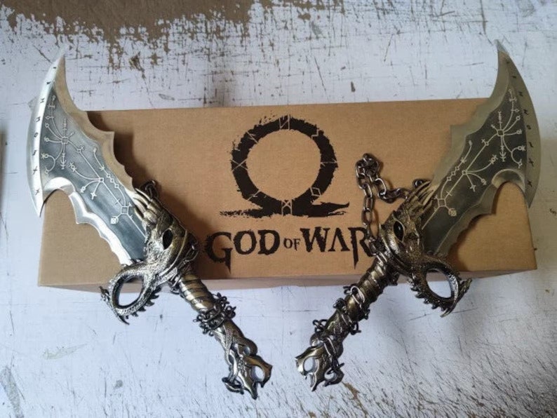 以下が含まれることがあります： 黒い柄のついた、銀色の装飾的な剣のペア。剣は、「God of War」と書かれた茶色の箱の上に置かれています。