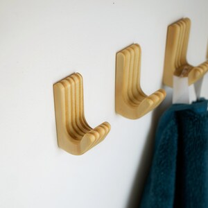 May include: Wooden wall hooks with a curved design, mounted on a white wall. The hooks are made of light-coloured wood and have a grooved texture. A teal towel hangs from one of the hooks.