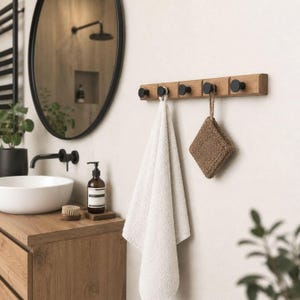 Op de afbeelding: Een houten handdoekenrek met vijf zwarte knoppen aan de muur. Een witte handdoek en een bruine washand hangen aan de haken. De badkamer heeft een ronde spiegel, een houten wastafelmeubel en een zwarte kraan.