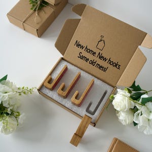 May include: A brown cardboard box containing four J-shaped wooden hooks with red accents, set in white foam. The inside lid features a house icon and the text: New home. New hooks. Same old mess! One additional wooden hook rests outside the box.