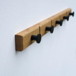 Op de afbeelding: Een houten kapstok met zes zwarte ronde haken. De kapstok is gemaakt van lichtbruin hout en is aan een witte muur bevestigd. De haken zijn gelijkmatig verdeeld over de lengte van de kapstok.