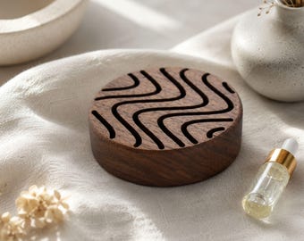May include: A circular dark brown wooden essential oil diffuser featuring a series of carved, wavy, parallel cutouts across its top surface. The wood grain is visible, and the object has a smooth, cylindrical side profile with a flat base.