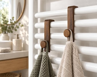 May include: Two dark brown wooden hooks with a curved top design are hanging over the horizontal bars of a white towel radiator. A sage green waffle-textured towel hangs from the left hook, and a beige terry cloth towel hangs from the right hook.
