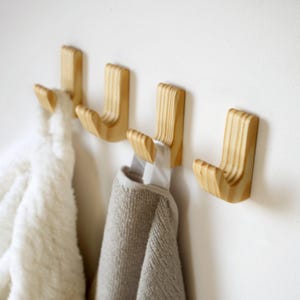 Puede incluir: Cuatro ganchos de pared de madera con un diseño curvo están montados en una pared blanca. Dos ganchos sostienen una toalla blanca y una toalla gris. Los ganchos son de madera clara con vetas visibles.