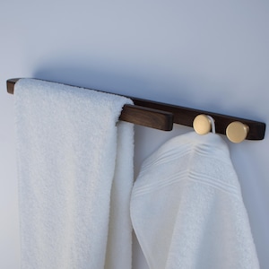 May include: A dark brown wooden towel rail with two gold-coloured knobs. A white towel and a white bathrobe hang from the rail. The rail is mounted on a white wall. The design is modern and minimalist.