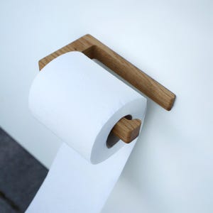Könnte beinhalten: Ein Toilettenpapierhalter aus Holz mit einer Rolle weißem Toilettenpapier. Der Halter ist aus hellbraunem Holz gefertigt und an einer weißen Wand montiert. Das Design ist schlicht und modern.