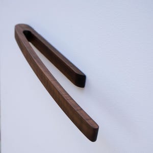 Könnte beinhalten: Ein dunkelbrauner Möbelgriff aus Holz mit einem glatten, länglichen und geschwungenen Design. Der Griff hat eine durchgehende Schlaufenform, die an einer flachen Oberfläche befestigt ist, und zeigt eine reiche Holzmaserung sowie ein poliertes Finish.