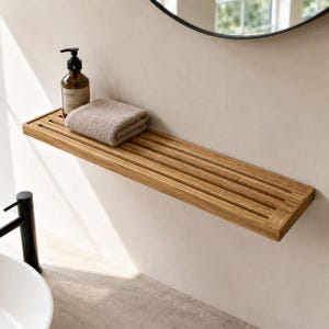 May include: A rectangular wall-mounted wooden shelf featuring a slatted design. A folded beige hand towel and a brown glass pump bottle with a black dispenser are placed on the surface of the shelf, which is mounted on a plain light-coloured wall.