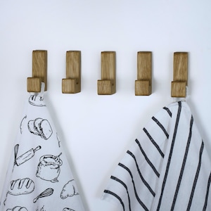 Peut inclure: Cinq crochets en bois montés sur un mur blanc. Deux torchons sont suspendus aux crochets. L'une des serviettes a un motif rayé noir et blanc. L'autre serviette a un motif noir et blanc de pain, un rouleau à pâtisserie, un fouet et un sac de farine.