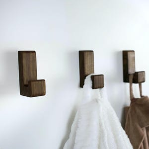 Può includere: Tre ganci in legno marrone scuro montati su una parete bianca. Due ganci hanno asciugamani bianchi e marroni appesi.