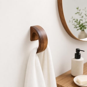 May include: A curved, dark brown wooden wall hook mounted on a white wall, holding a white fabric towel. The hook has a smooth, polished finish and a semi-circular shape, providing a functional storage solution for hanging bathroom linens.
