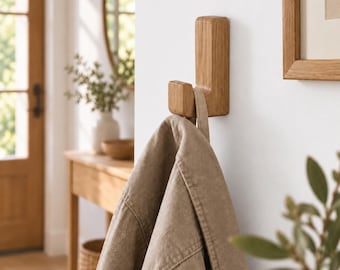 May include: A light brown wooden wall hook with a rectangular base and a curved upward-facing prong. A beige fabric jacket is hanging from the hook, showing the collar and shoulder area of the garment against a plain white wall surface.