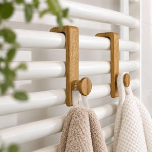 May include: Two light brown wooden hooks designed to hang over the horizontal bars of a white towel radiator. Each hook features a flat vertical body with a rounded knob at the bottom, currently holding a beige towel and a white textured towel.