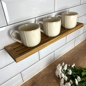Puede incluir: Tres tazas de cerámica blanca se colocan en un estante de madera con ranuras. El estante está montado en una pared de azulejos blancos.