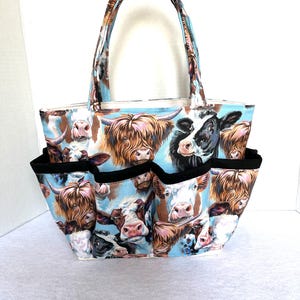 May include: A fabric tote bag with a cow print design. The bag features multiple pockets with black trim and a matching handle. The cow print includes various cow faces on a light blue background.