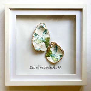 May include: Framed artwork with two oyster shells, each decorated with a butterfly design in blue and green. The shells have gold edges and are set against a white background. The text "Wild and free just like the sea" is written below the shells.