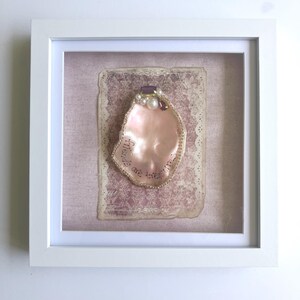 May include: A square white shadow box frame containing a pink oyster shell decorated with white pearls and purple crystals. The shell features the handwritten text "Friends are treasures" in black ink, mounted on a vintage-style patterned paper.
