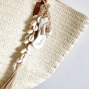 May include: A woven straw tote bag with brown leather handles. A seashell and rope keychain with the text "little things" hangs from the bag. The keychain features a collection of white seashells and a tassel.
