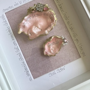 May include: A white shadow box frame containing two pink-painted oyster shells with gold-painted edges. The larger shell features the text 'chase your dreams and catch them' and a rhinestone accent. The smaller shell features the word 'friends' and pearl accents. The background mat includes the text 'Someone who is by your side throughout all of lifes dramas, laughs and cries with you. I love you' and 'an irreplacable friend that you immediately click with' and 'Soul Sister'.