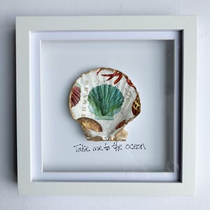 May include: A white framed artwork with a seashell design. The shell is decorated with teal and gold accents, red starfish, and the words "SEASHELL" and "Take me to the ocean". The artwork is a decorative piece.