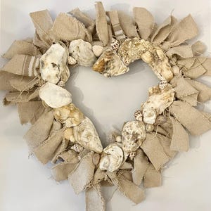 Peut inclure: Couronne en forme de cœur composée de ruban de jute naturel, de coquilles d'huîtres et de petits coquillages. La couronne est de couleur neutre, avec les coquillages et la toile de jute créant un thème côtier rustique.
