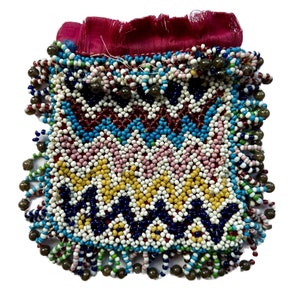 Könnte beinhalten: Eine kleine, geperlte Tasche mit einem roten Stoffdeckel und einem Fransenbesatz aus Perlen. Die Tasche ist mit einem geometrischen Muster aus blauen, weißen, rosa, gelben und roten Perlen verziert.