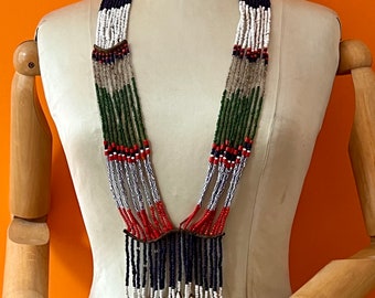 May include: A multi-strand beaded necklace with green, white, red, and blue beads. The necklace has a fringe of blue beads and white shell accents.