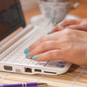 May include: A white laptop computer with a black keyboard and a silver touchpad. The laptop is open and a person's hands are typing on the keyboard. The person's fingernails are painted a light blue color.