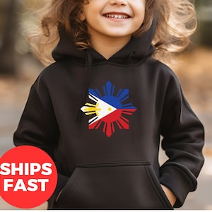May include: A black hoodie with a large white sun design featuring the Philippine flag colors. The sun has 8 rays and the flag is in the center. A young child is wearing the hoodie and smiling.
