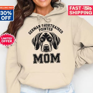 May include: A beige hoodie featuring a black and white graphic of a German Shorthaired Pointer dog's face, with the word "MOM" below. The text "GERMAN SHORTHAIRED POINTER" arches above the dog. The hoodie has a drawstring hood.