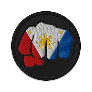 Peut inclure: Écusson brodé représentant un poing serré. Le poing est aux couleurs du drapeau philippin : rouge, blanc et bleu. Un soleil et des étoiles jaunes sont au centre. L'écusson a une bordure noire.