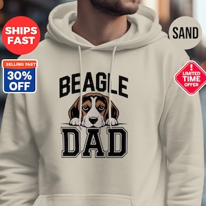 Könnte beinhalten: Ein sandfarbener Kapuzenpullover mit dem Schriftzug "BEAGLE DAD" in Schwarz und einer Beagle-Grafik. Der Hoodie hat eine Kapuze mit Kordelzug und eine Vordertasche. Werbeaufkleber sind ebenfalls zu sehen.