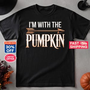 May include: A black short-sleeved t-shirt featuring the white text I'M WITH THE PUMPKIN centered on the chest. Between the two lines of text is a horizontal arrow graphic in a light orange color. The shirt has a crew neckline and a standard fit.
