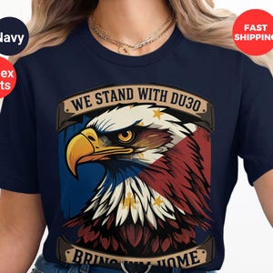 May include: A navy blue t-shirt featuring a graphic of a bald eagle with the Philippine flag on its chest. The text "WE STAND WITH DU30. BRING MY HOME" is printed on the shirt.
