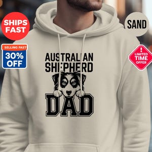 May include: A cream-coloured hoodie featuring the text "AUSTRALIAN SHEPHERD DAD" in black, with a graphic of an Australian Shepherd dog. The hoodie has a hood and a front pocket.