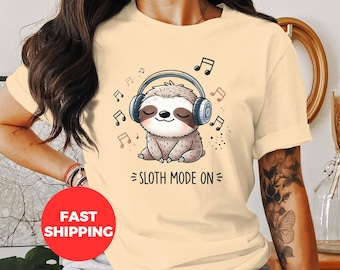 Könnte beinhalten: Ein hellbeiges T-Shirt mit einem Cartoon-Faultier, das Kopfhörer trägt und den Text "Sloth Mode On".
