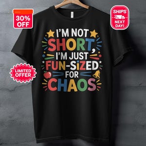 May include: Black t-shirt with the text "I'M NOT SHORT, I'M JUST FUN-SIZED FOR CHAOS" in colourful letters. The design includes stars, an apple, and a bell. The shirt is hanging on a wooden hanger.