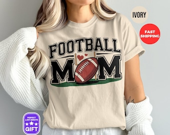 Op de afbeelding: Een beige T-shirt met de tekst "FOOTBALL MOM" in zwarte letters. Onder de tekst zijn een voetbal en een doelpaal afgebeeld. Het shirt is lichtbeige.