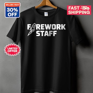 May include: A black short-sleeved t-shirt featuring white text that reads FIREWORK STAFF, with the letter I replaced by a graphic of a rocket ship launching upwards. The shirt is displayed on a wooden hanger against a neutral gray background.