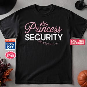 May include: A black short-sleeved t-shirt featuring the text Princess in pink script with a crown icon above it, followed by the word SECURITY in bold white capital letters, with a trail of small pink dots swirling beneath the text.
