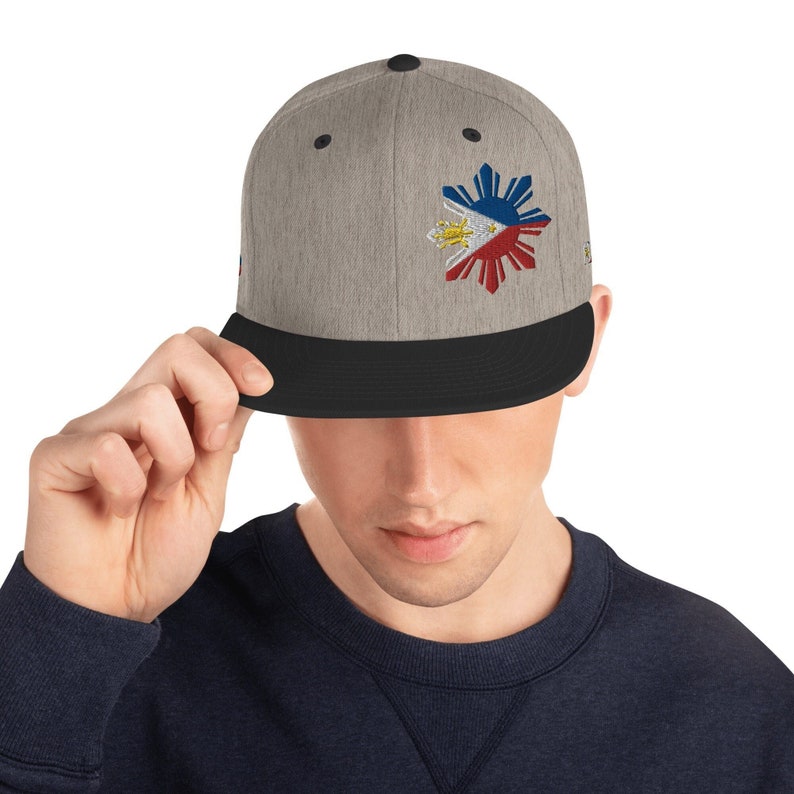 Pu&ograve; includere: Un cappello da baseball grigio e nero con una bandiera filippina ricamata sul davanti. La bandiera &egrave; rossa, bianca e blu con un sole giallo al centro.