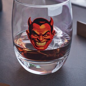 May include: A clear glass with a red devil face graphic on the side. The glass is filled with a dark liquid and ice cubes.